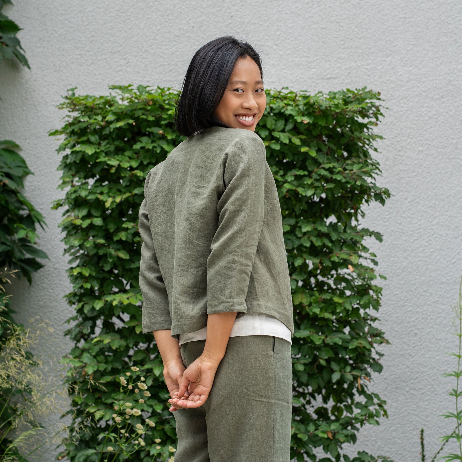 A woman wearing a white top, gray jacket, and gray pants poses outdoors against a green leafy background.