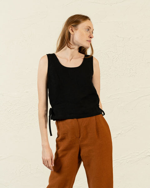 A studio shot of a woman with long, light brown hair looking to her right. She is wearing a black sleeveless top with a scoop neck and side ties, paired with high-waisted, rust-colored pleated trousers. Her arms are down at her sides. The background is a plain, light-colored wall.
