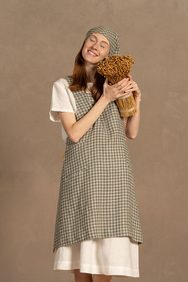 A person with fair skin and long reddish-brown hair stands smiling with closed eyes, gently holding a bundle of dried golden flowers to their chest. They are wearing a white short-sleeved linen shirt under a green check apron and a matching checkered headscarf tied under their chin. The background is a muted warm brown.