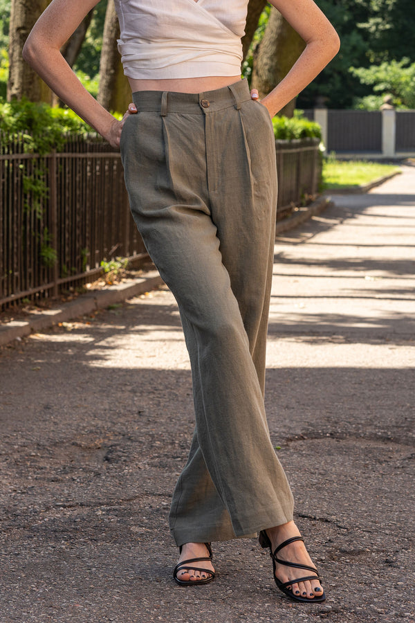 A person is shown from the waist down, modeling stone green, high-waisted, linen wide-leg trousers. They are wearing black open-toe sandals with thin straps and have their hands on their hips. The setting appears to be outdoors on an asphalt path, with a black metal fence and greenery visible in the background.