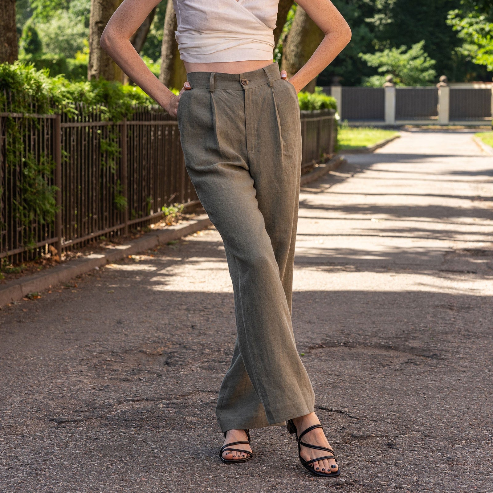 A fair-skinned woman models a cinnamon red colored linen vest and wide-leg trousers, standing against a textured light beige wall. She is wearing black sandals.