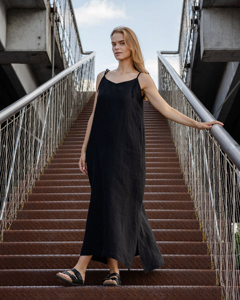 A light-skinned person with long blonde hair is standing on a set of metal stairs with a walkway above. They are wearing a long black menique sleeveless dress with thin straps and dark sandals, and are holding onto a metal railing with one hand. The background shows a somewhat overcast sky.