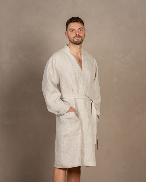 A man, turned slightly to the side, wearing the men's linen bathrobe in natural color. Both of his hands are in the side pockets.