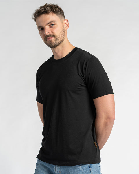 A man wearing a merino wool black t-shirt and blue jeans is shown from the chest up against a light background.  He has short brown hair and a light beard, and is looking slightly to the side. The menique black t-shirt is the primary focus, showcasing its fit and appearance.