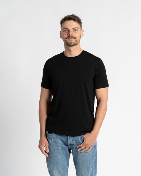 A man with short, dark brown hair and a neatly trimmed beard stands centered against a plain white background, directly facing the viewer. He wears a black crew neck t-shirt with short sleeves from menique brand, showcasing the garment's fit and simplicity. The t-shirt is made of merino wool fabric. He is also wearing light blue denim jeans with a straight leg cut and a visible front pocket, with one hand casually tucked into the pocket. 