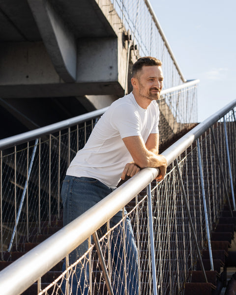 A man with a light beard stands on an outdoor staircase, leaning casually with both arms on a metal handrail and looking off to the right. He is wearing a fitted white short-sleeve t-shirt and blue jeans. The background features a concrete structure and wire mesh fencing.