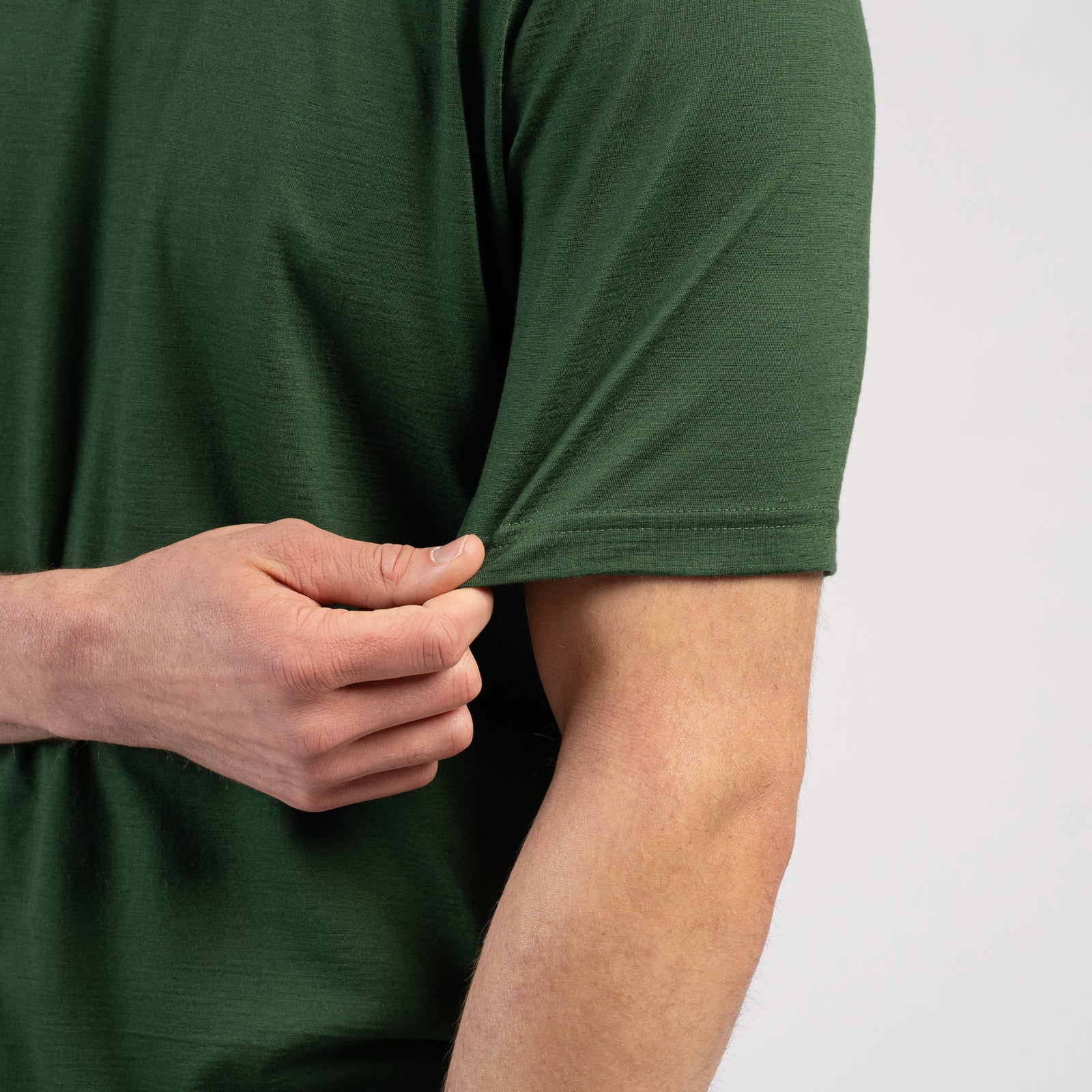 A studio portrait of a man standing against a plain white background. He is looking off to the right side of the frame. He has medium-length brown hair and a light beard. He is wearing a short-sleeved, dark green crew-neck t-shirt with a regular fit, paired with medium-wash blue jeans. His arms are relaxed by his sides. There is a small tag visible on the lower left hem of the t-shirt.