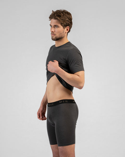 A studio portrait of a man standing in profile facing left, looking straight ahead against a plain light grey background. He is wearing a charcoal grey short-sleeved t-shirt and matching long boxer briefs, gently lifting the front of his t-shirt with one hand to show his stomach and the waistband.