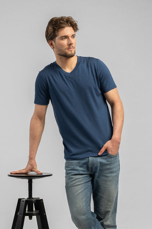 A man stands leaning casually with his right hand on a black stool, looking off to the right. He wears a denim blue v-neck t-shirt and blue jeans against a light grey background.