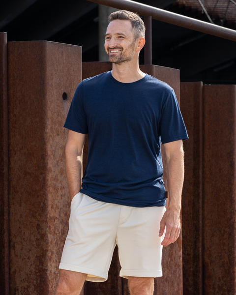 A smiling man standing outdoors in front of rusty metal pillars. He is wearing a navy blue short-sleeve crew neck t-shirt and beige shorts, with his right hand casually resting in his pocket as he looks off into the distance.