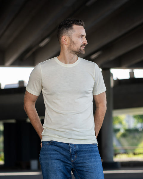 A man standing outdoors under a concrete overpass, looking away to his left. He is wearing a natural, off-white short-sleeve crew neck t-shirt and blue jeans, with his hands tucked into his back pockets.