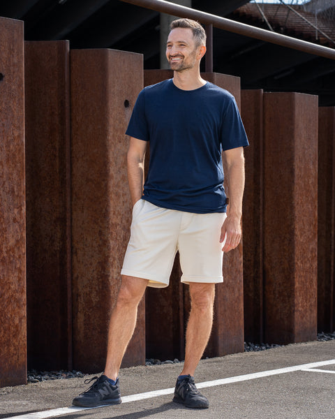 A full-body shot of a smiling man standing on an asphalt surface next to a row of rusty metal pillars. He is wearing a short-sleeve navy blue crew neck t-shirt, beige shorts, and dark sneakers, with his right hand casually in his pocket.