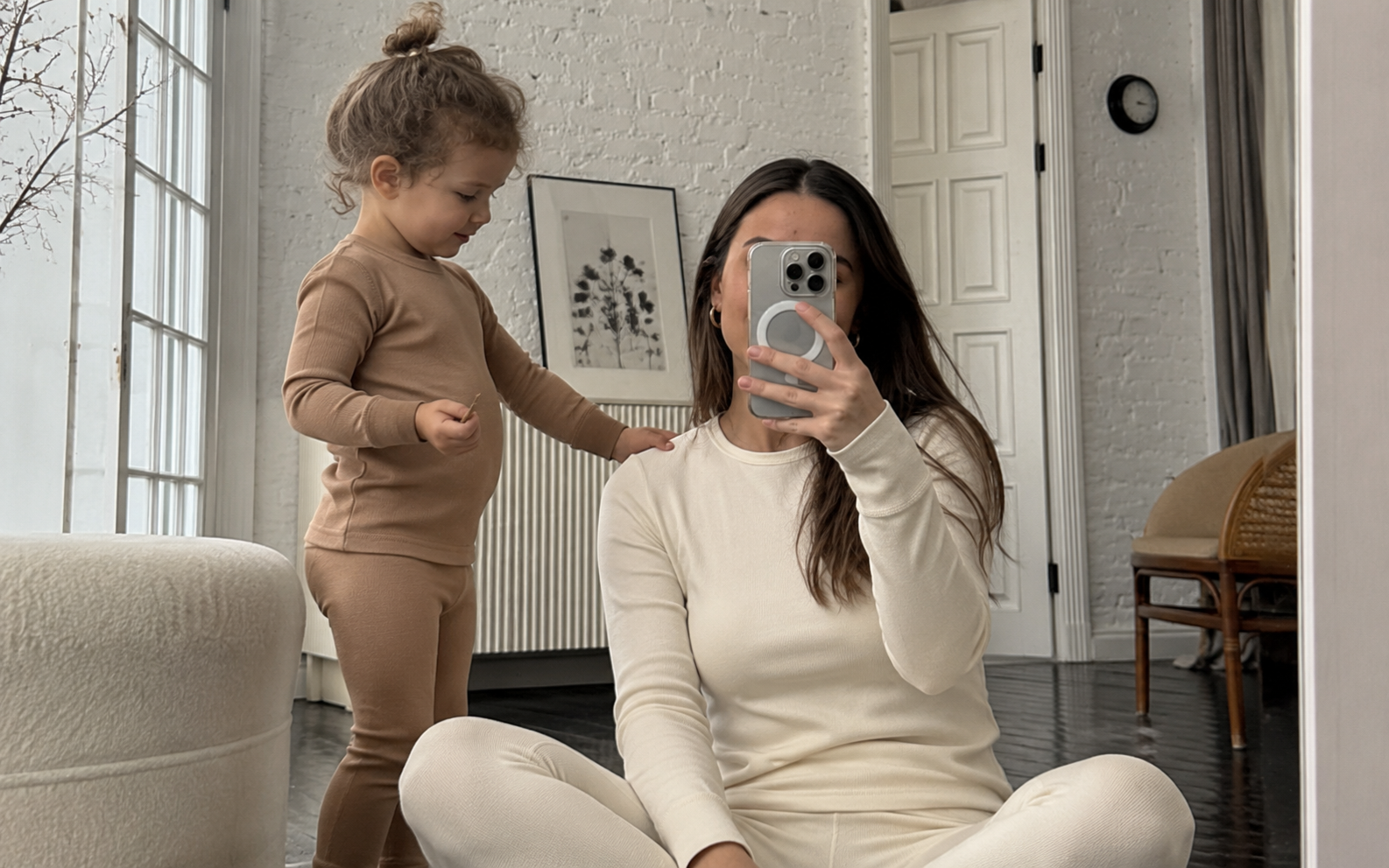 A woman sitting on the floor taking a mirror selfie, wearing a cream-colored ribbed thermal set, with a toddler standing next to her in a matching light brown set.