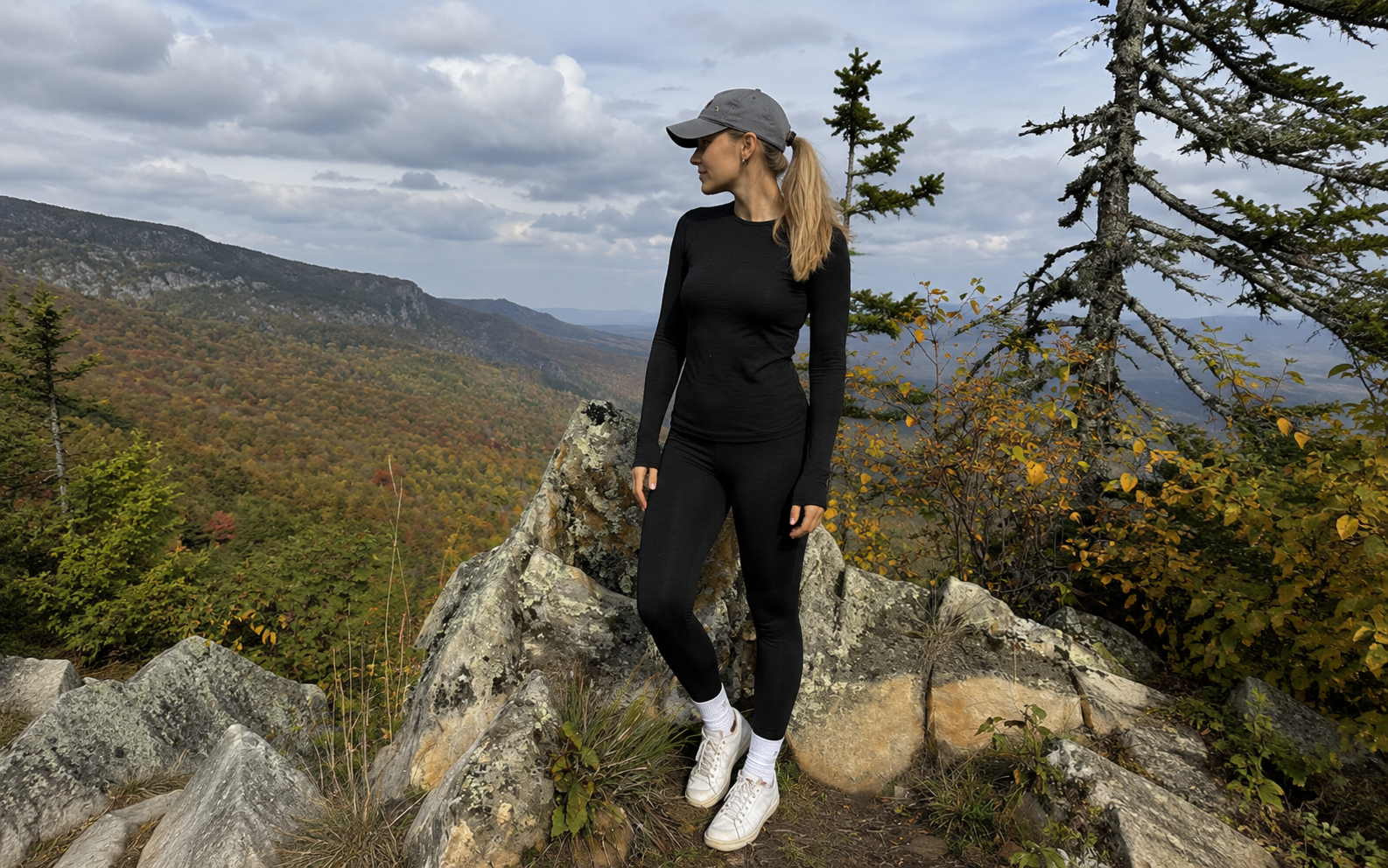 A woman standing on a rocky mountain overlook with autumn trees in the background, wearing a fitted black long-sleeved top, black leggings, white sneakers, and a grey baseball cap.