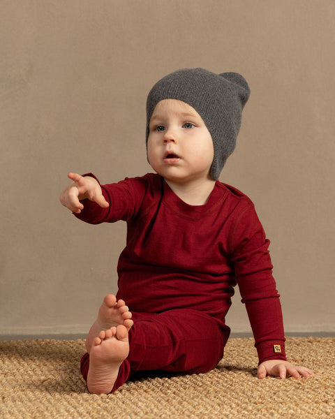 Baby wearing a merino wool royal cherry outfit and gray beanie sitting on a textured surface with a beige background