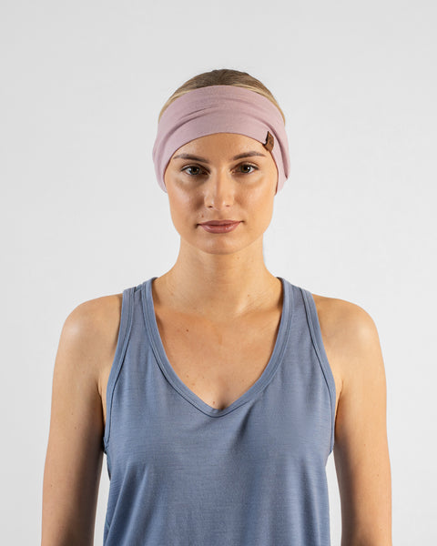 A studio portrait of a blonde woman facing forward and looking directly at the camera. She is wearing a dusty blue v-neck sleeveless top and a wide dusty pink headband with a small brown logo tag against a plain light grey background.