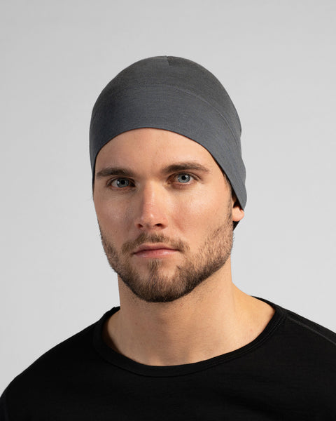 A close-up studio portrait of a man looking directly at the camera. He is wearing a fitted, medium grey skull cap and a solid black crew-neck top. The background is a plain light grey.