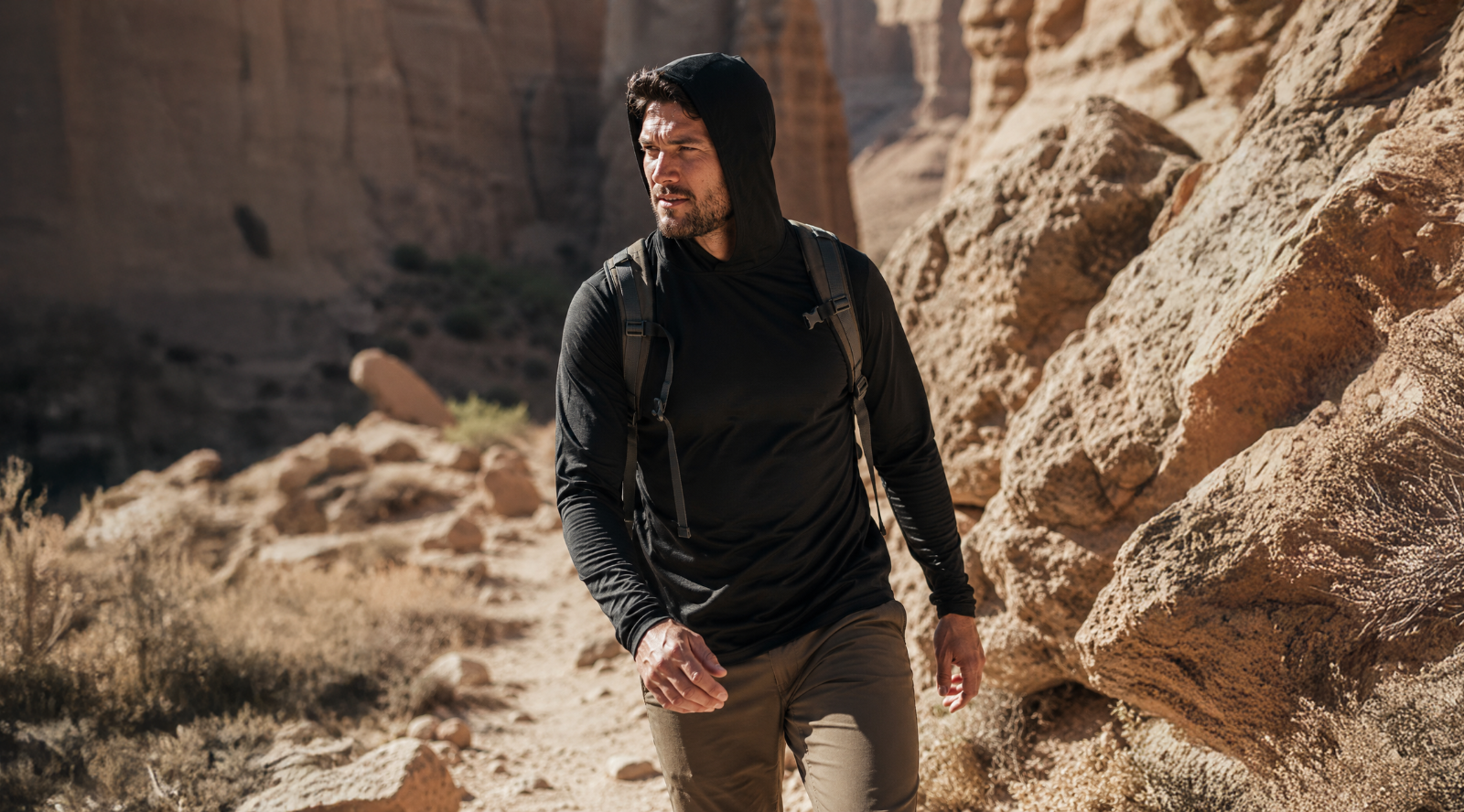 An outdoor shot of a man hiking along a rugged, sunlit canyon trail. He is looking off to the left side of the frame, wearing a black long-sleeved hooded shirt with the hood pulled up, olive green pants, and a backpack.