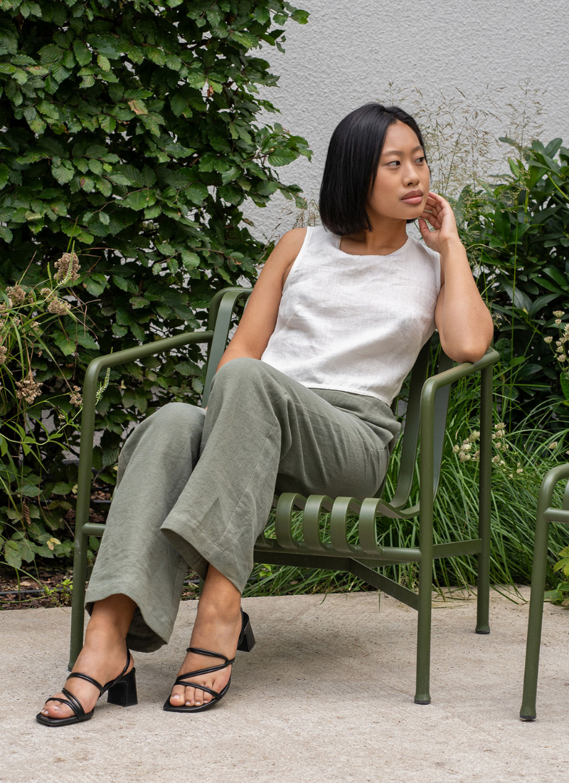 A woman with short black hair is sitting on a green metal chair in a garden, wearing a sleeveless white linen top and relaxed-fit olive green linen trousers. She is looking to the side with a calm expression, surrounded by lush greenery and plants, creating a peaceful outdoor setting.