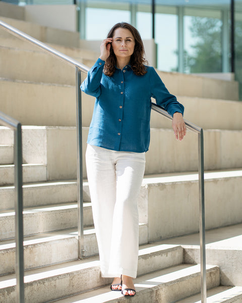 A woman with brown hair, wearing a cobalt blue button-front shirt and wide-leg white trousers, stands outdoors against a modern concrete structure.