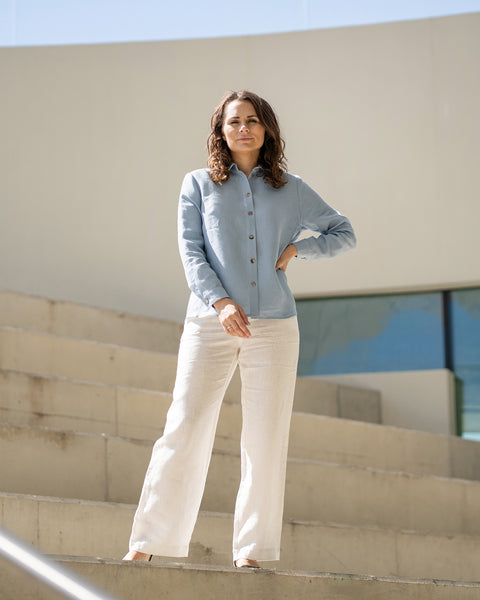 A woman with brown hair, wearing a light blue button-front shirt and wide-leg white trousers, stands outdoors against a modern concrete structure.

