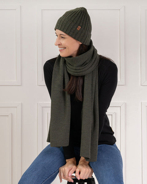 A smiling woman sitting down, looking off to the side. She is wearing a black long-sleeved top, blue jeans, and a matching olive green knitted winter set consisting of a ribbed beanie with a small brown leather logo patch and a long scarf wrapped around her neck. The background is a bright white wall with classic decorative paneling.