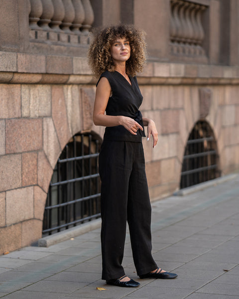 A woman with a voluminous mass of light brown, curly hair stands on a sidewalk, looking towards her left. She is wearing a black linen sleeveless wrap top with a V-neck and black wide-leg trousers, completed with black flat shoes. 
