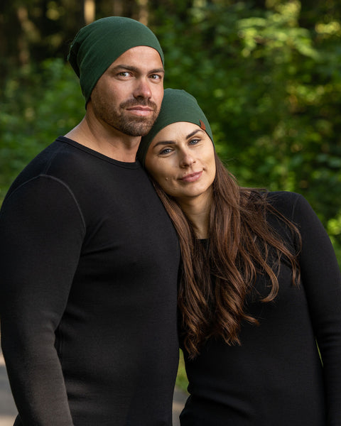 A half body upper body picture of a couple wearing unisex dark green menique merino headbands. They are smiling, the woman is leaning on the man's shoulder. They are also wearing black long sleeve merino tops.