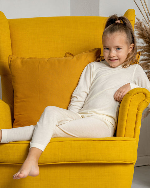 A girl sitting in an armchair in yellow color, wearing the menique kids' pants from merino fabric in natural color. 