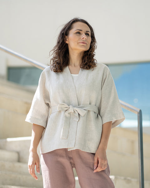 A woman with brown hair, wearing a light beige kimono-style jacket over mauve trousers, stands on indoor concrete stairs.
