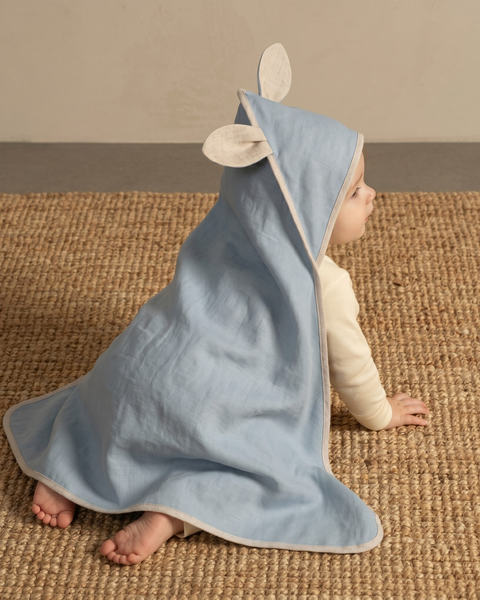 A baby crawling on a woven natural fiber rug, viewed from the side and slightly behind. The baby is wearing a light dusty blue hooded baby towel over a light-colored long-sleeve top. The hood is pulled up, featuring two small, cute animal-like ears made of a contrasting light beige fabric, which matches the binding along the edges of the towel. The baby's bare feet are visible. The background is a solid, warm taupe wall.