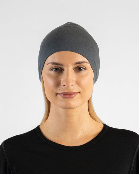 A studio portrait of a woman looking directly at the camera. She is wearing a fitted, medium grey skull cap over her blonde hair and a black top. The background is a solid light grey.