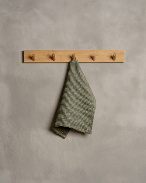 A gray wall with a wooden hanger. In the middle of it hangs a waffle face washcloth in stone green color.
