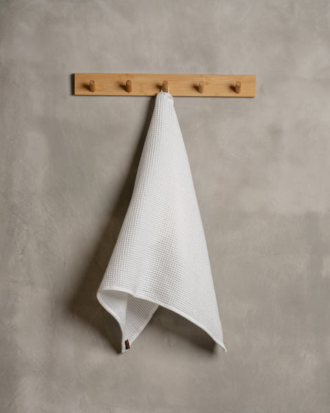 A gray wall with a wooden hanger. In the middle of it hangs a waffle hand washcloth in pure white color.