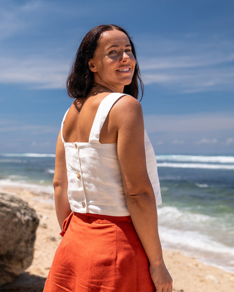 A woman with shoulder-length dark hair, wearing a pure white linen sleeveless crop top with button closures down the back and menique linen skirt, stands on a sandy beach looking over her left shoulder towards the ocean under a bright sky with scattered clouds.