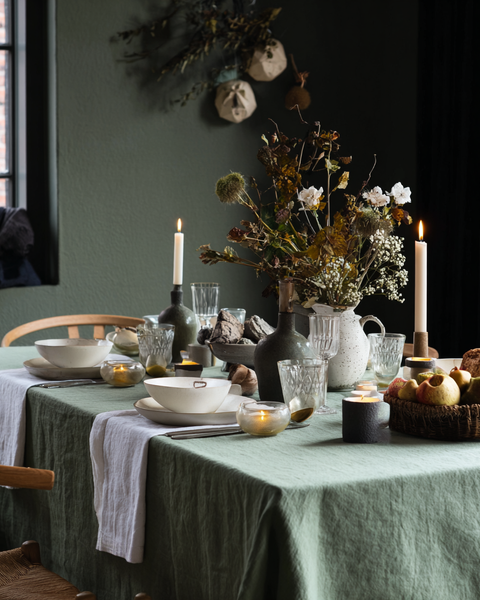 Cozy fall dining table decorated with green linen tablecloth, white linen napkins, candles, and natural rustic centerpiece for holiday hosting.