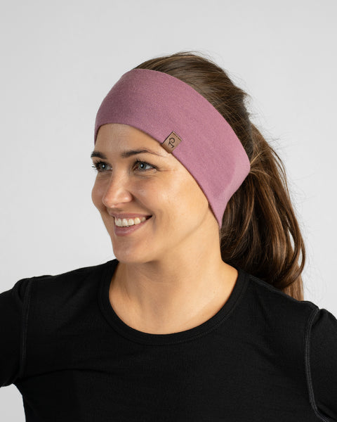 A smiling woman looking to the side against a neutral backdrop, wearing a dusty rose-pink knit headband with a black long-sleeve top.