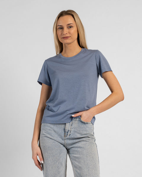 A studio portrait of a blonde woman standing and looking directly at the camera, with her left hand casually tucked into the front pocket of her light-wash blue jeans. She is wearing a regular fit, dusty blue short-sleeved crew-neck t-shirt against a solid light grey background.