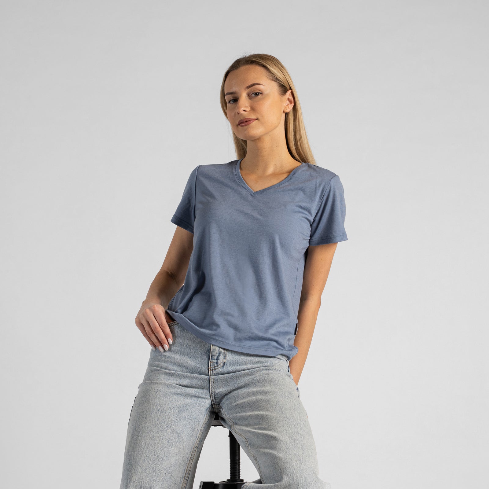 A studio portrait of a blonde woman sitting on a stool and looking directly at the camera. She is leaning back slightly, with her hands resting on her lap. She is wearing a regular fit, dusty blue short-sleeved v-neck t-shirt and light-wash blue jeans against a plain light grey background.