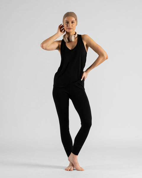 A woman kneeling barefoot on a white background, wearing a black sleeveless  top, black leggings, and a beige headband. Beige headphones rest around her neck as she poses with one hand on her hip, looking at the camera.