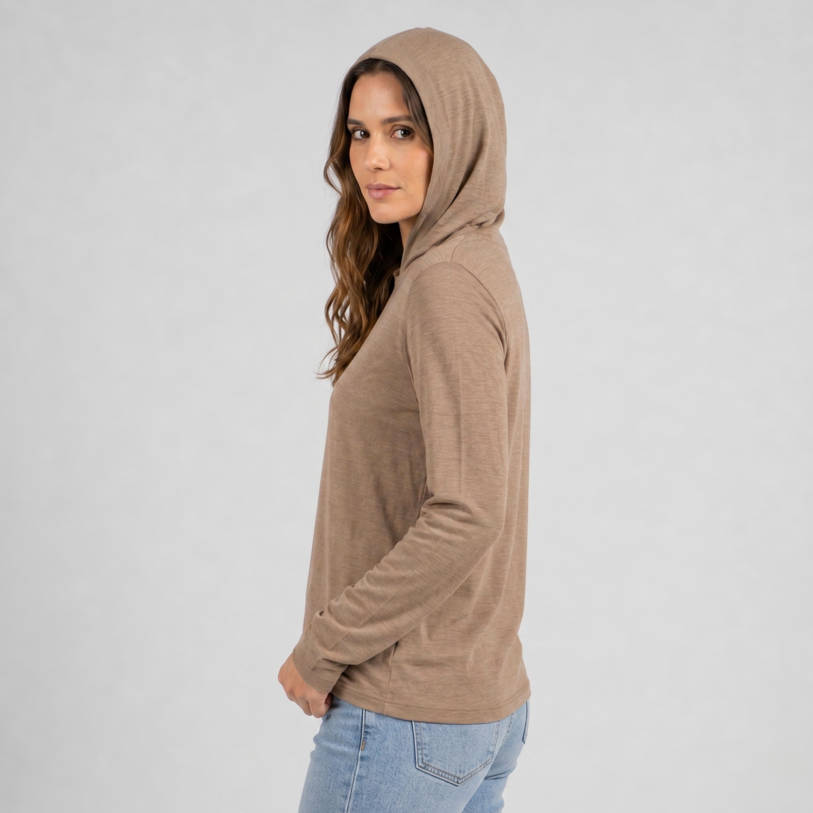 An outdoor portrait of a woman standing on a sandy path within a rocky canyon under a clear blue sky. She is looking directly at the camera with a gentle smile, wearing a lightweight beige long-sleeved hoodie with the hood pulled up and black pants, with one hand resting near the edge of the hood.