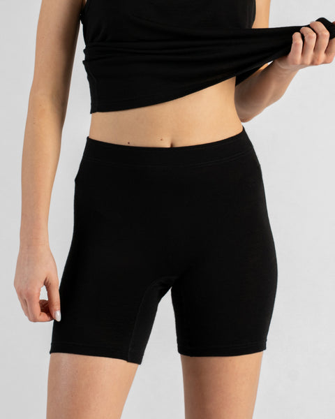 A model facing forward wearing black, form-fitting short leggings and a black top against a plain light background. Their left hand is lifting the hem of the top slightly, revealing their midriff, while their right arm hangs relaxed by their side.
