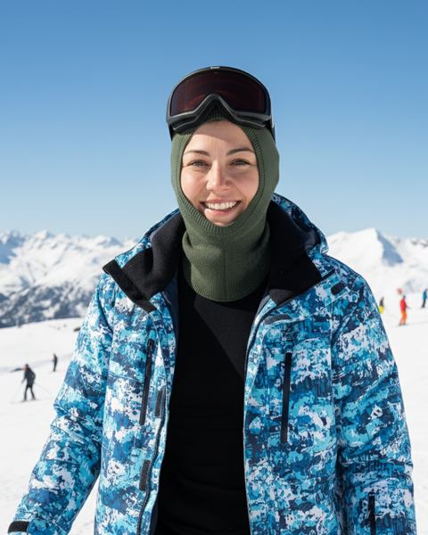 Smiling woman on ski slopes wearing a dark green Merino wool liner balaclava under a blue ski jacket and goggles.