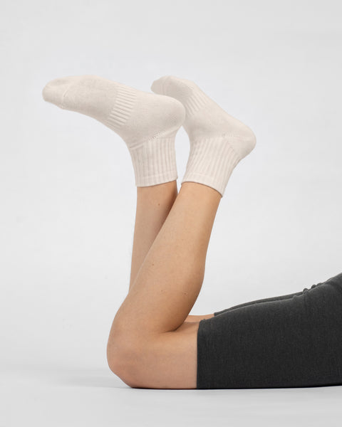 A person lying down with their knees bent and legs raised, wearing dark grey shorts and natural off-white crew socks against a plain white background.