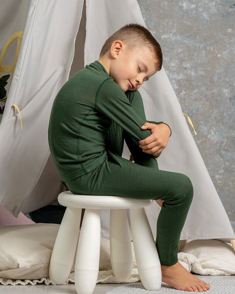 Child in dark green merino wool pajamas sitting on a white stool in a cozy indoor setting with a teepee.