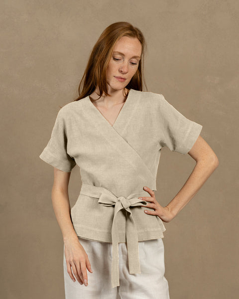 A fair-skinned person with long, auburn hair, wearing natural V-neck linen wrap top with short, wide sleeves and light-colored pants. They are holding the ends of the top's tie in front of them, forming a bow.