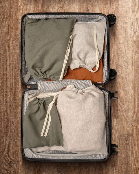An open suitcase is neatly packed with four cloth drawstring bags of different sizes and colors (two olive green and two light beige) on a wooden surface.