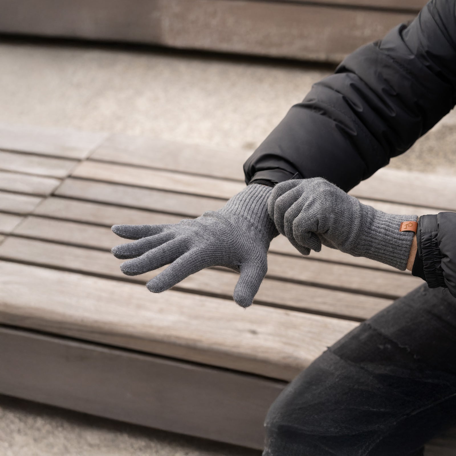 A person wearing black knit gloves from menique, while sitting on a wooden bench outdoors. The gloves appear to be snug-fitting and warm.