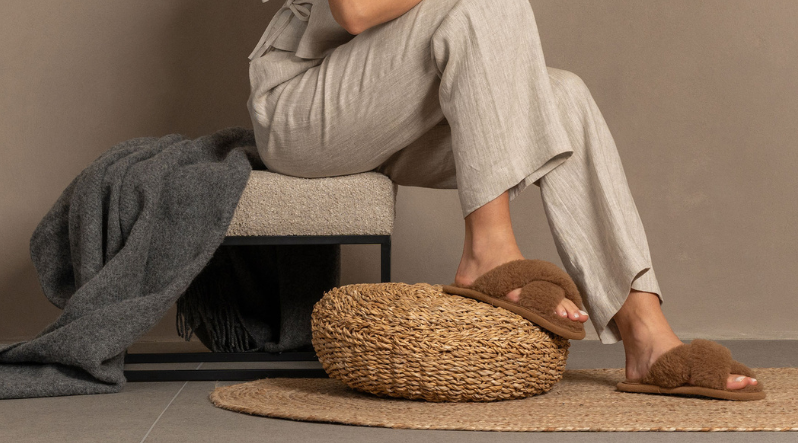 A person sits in a chair wearing light grey loungewear with their feet resting on a round, woven straw pouf.