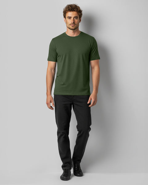 A man stands facing forward wearing a green T-shirt, black jeans, and black sneakers against a plain gray background.